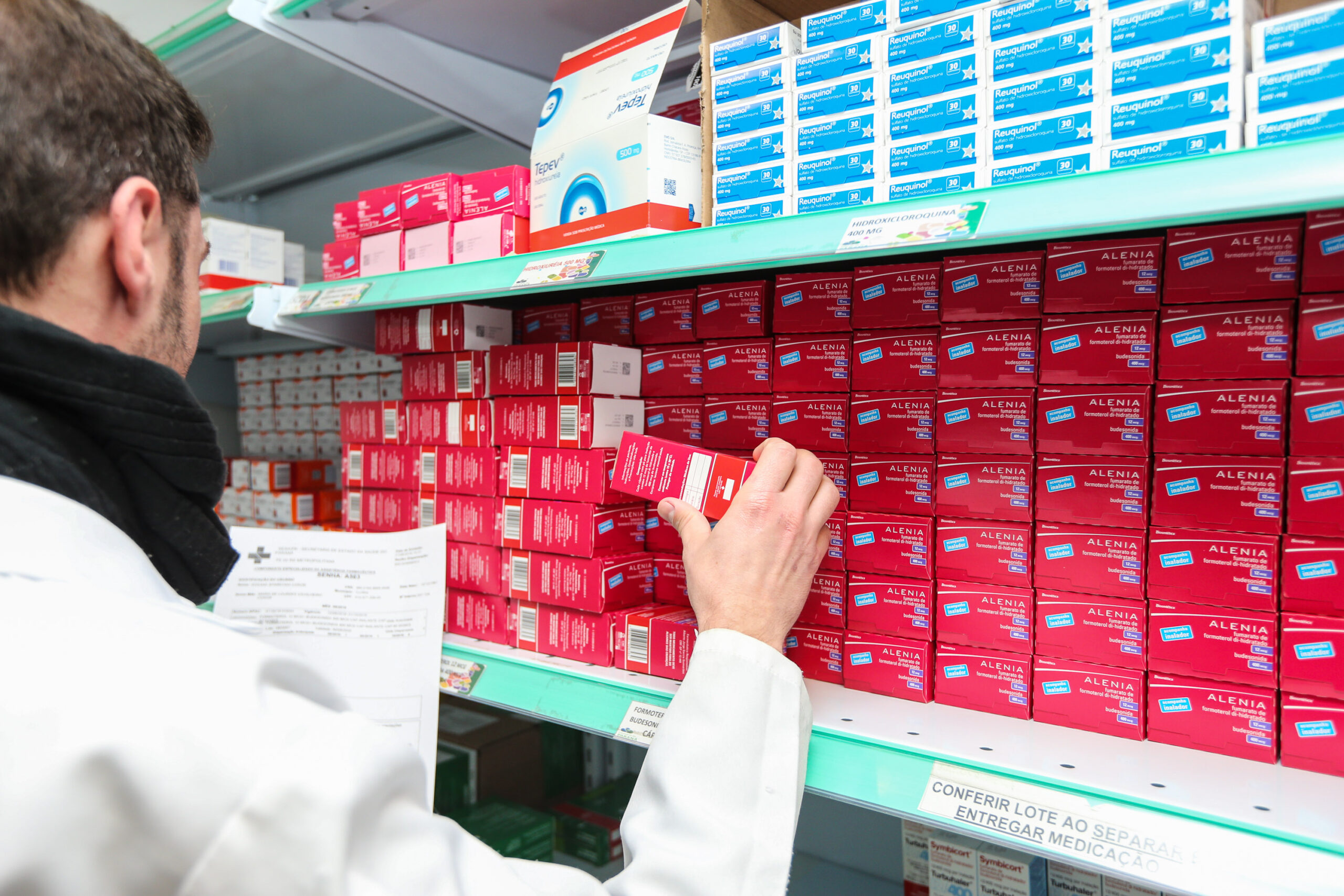Profissional de jaleco confere caixas de medicamentos em prateleiras de uma farmácia da rede pública do Paraná.